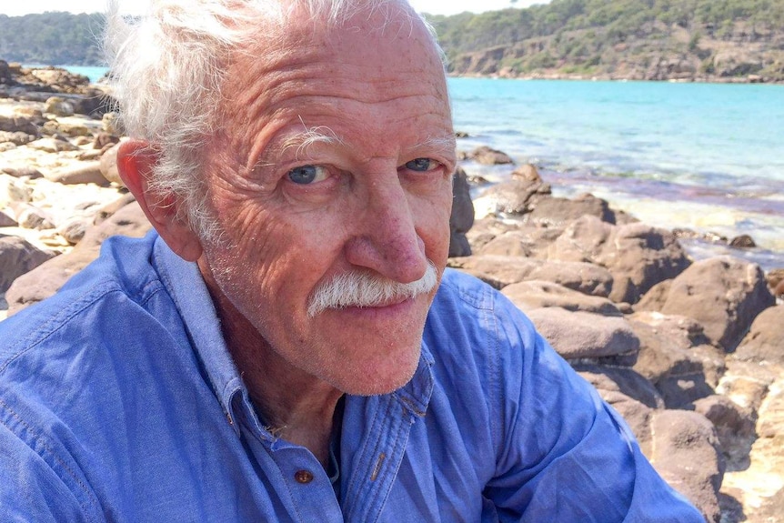 A man with blue eyes and a blue shirt stares down the lens, behind him is the turquoise water where a river meets the sea.