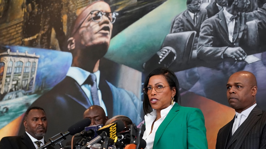 A middle-aged black woman in a green jacket and glasses speaks at a microphone-filled lectern in front of a Malcolm X mural.