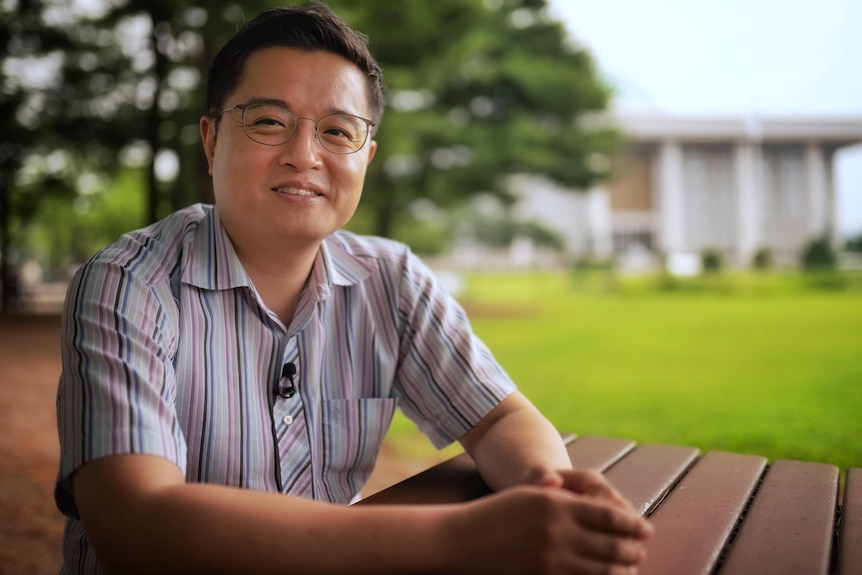 A man wearing a striped button up shirt and glasses smiles while sitting in a park.