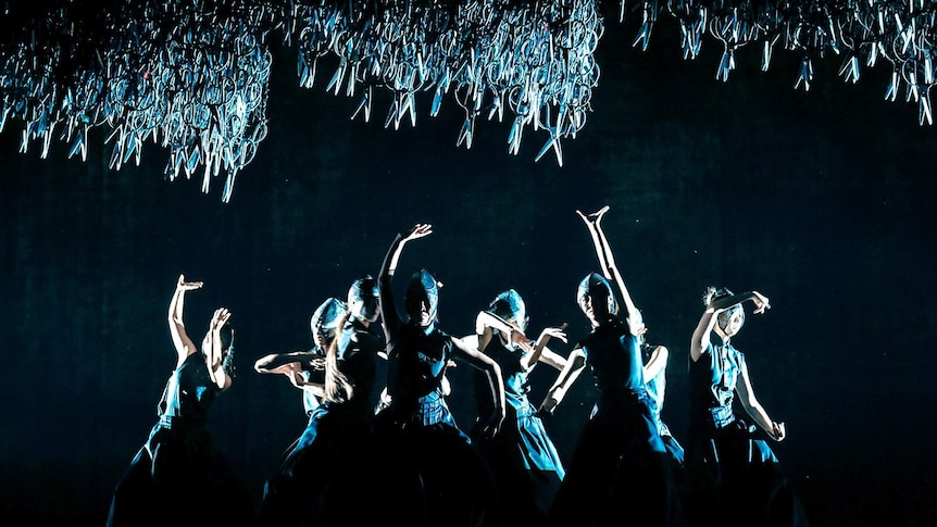 Scissors dangle above a group of dancers dressed in black.