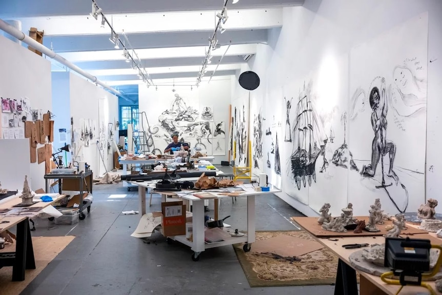 A white studio with large black drawings pinned on the walls and sculptures scattered, a woman working in the back