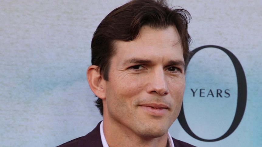 Ashton Kutcher stands against a light blue backdrop smiling slightly