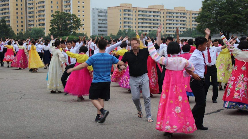 A pair of tourists dance among a group of people in North Korean traditional dress. One of the two men is wearing thongs.