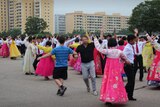 A pair of tourists dance among a group of people in North Korean traditional dress. One of the two men is wearing thongs.