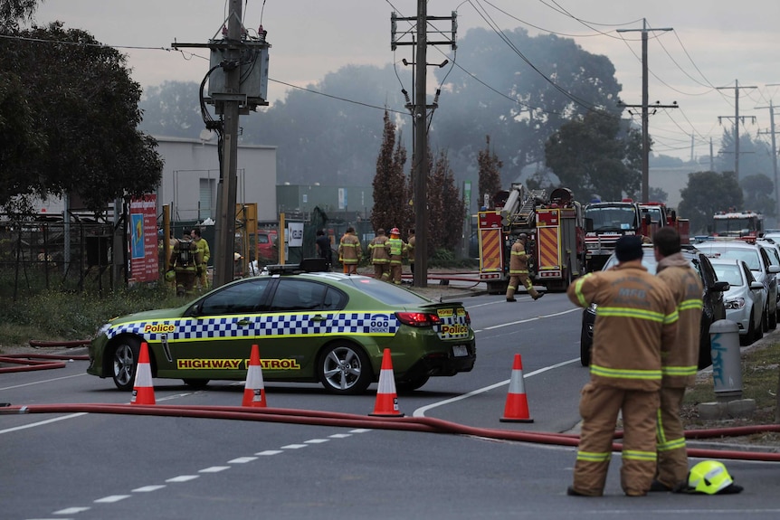 A police car blocks a road as firefighters gather outside an industrial site.