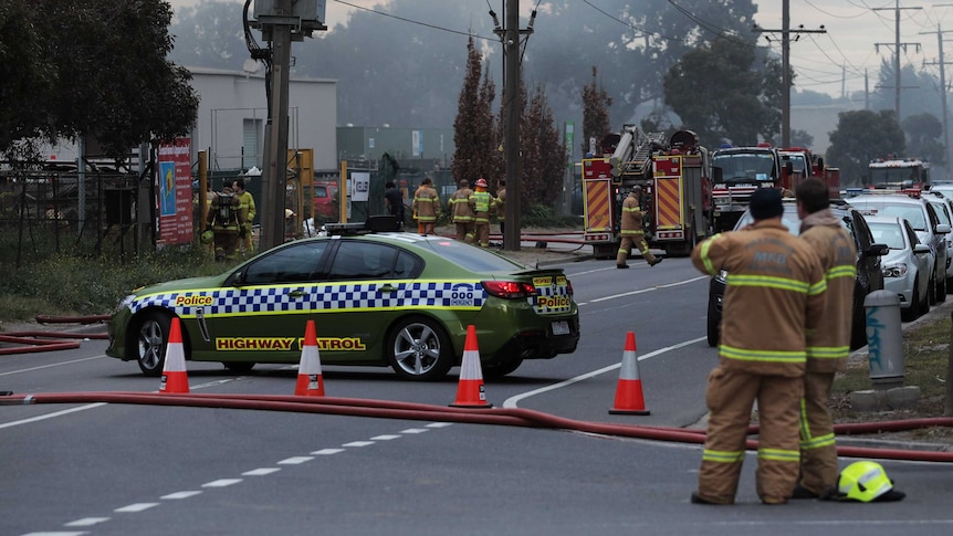 A police car blocks a road as firefighters gather outside an industrial site.