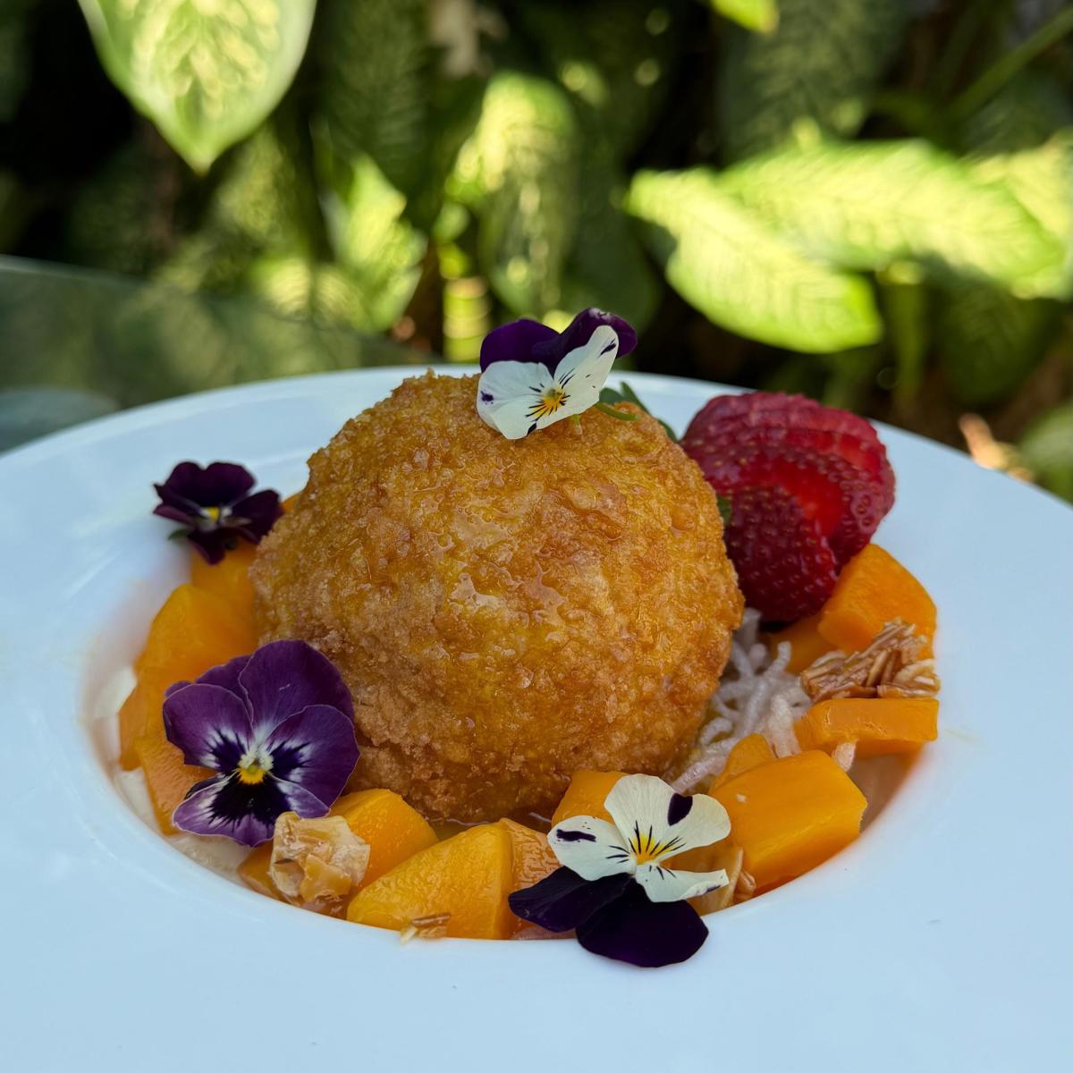 A large deep-fried scoop of ice cream sitting in a bowl, fresh fruit and small floral garnishes decorate the dish.