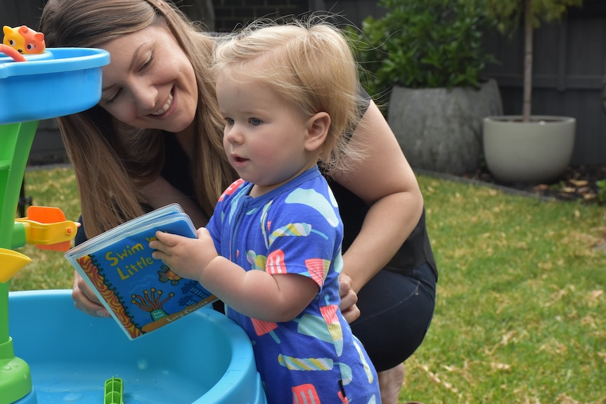 A woman with brown hair smiles as she plays with a baby in a backyard.