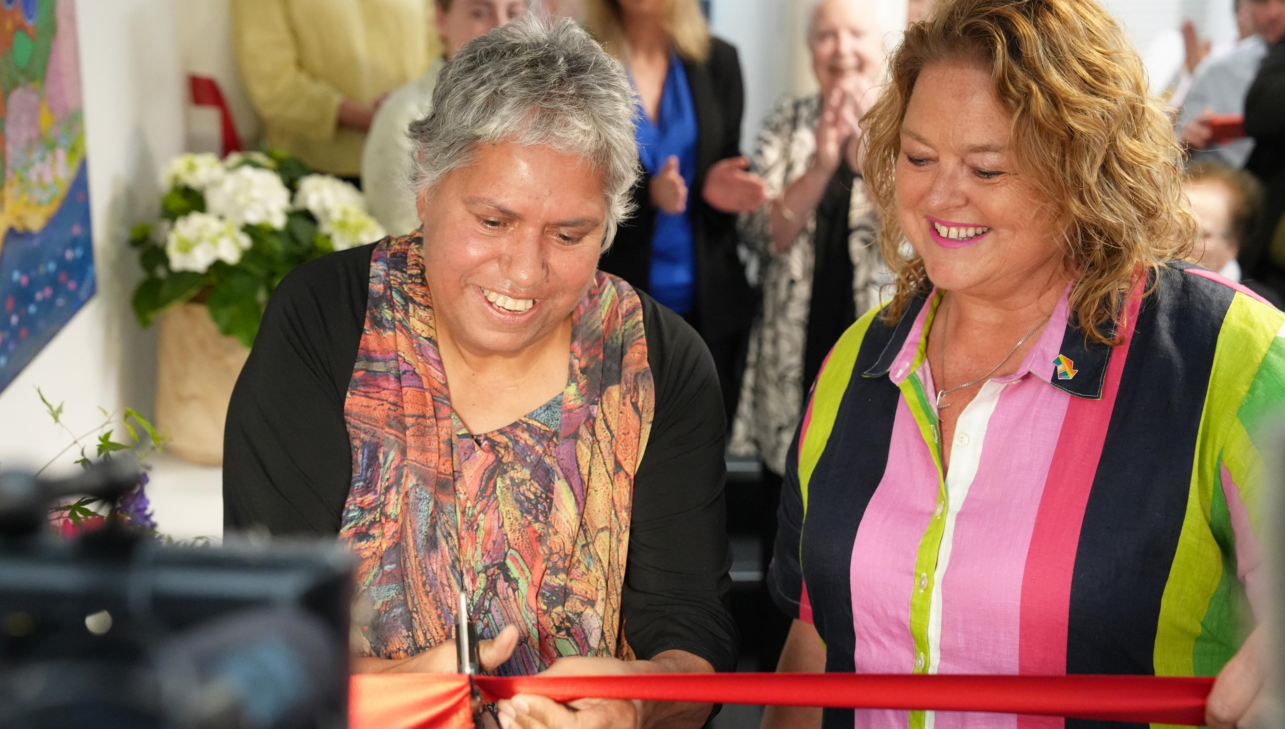 Two women smiling while one cuts a red ribbon.