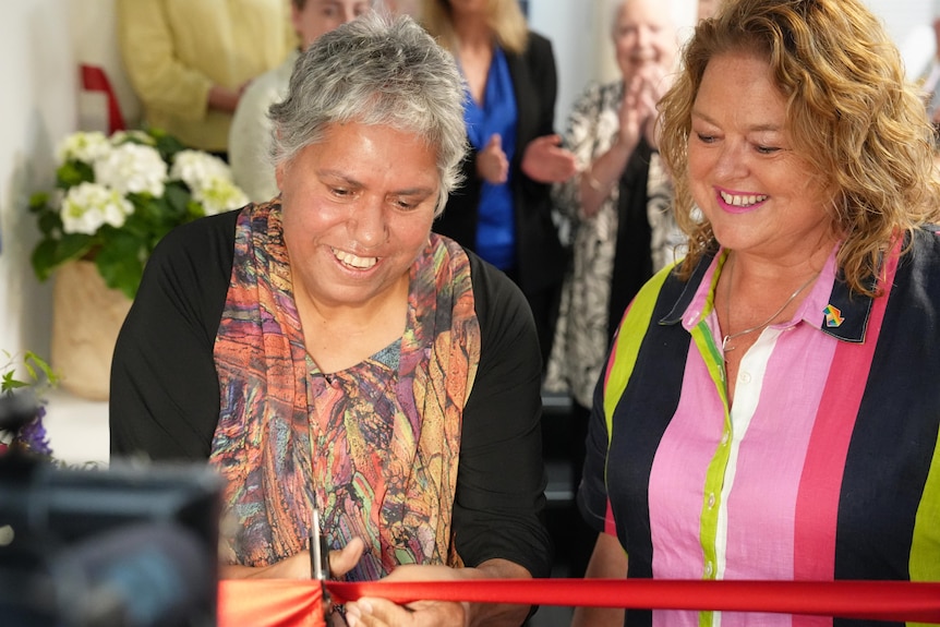 Two women smiling while one cuts a red ribbon.