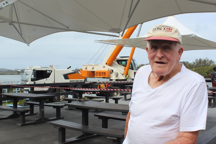 An elderly man with a cap on, standing in front of a set of tables. An orange and white crane can be seen in the background.