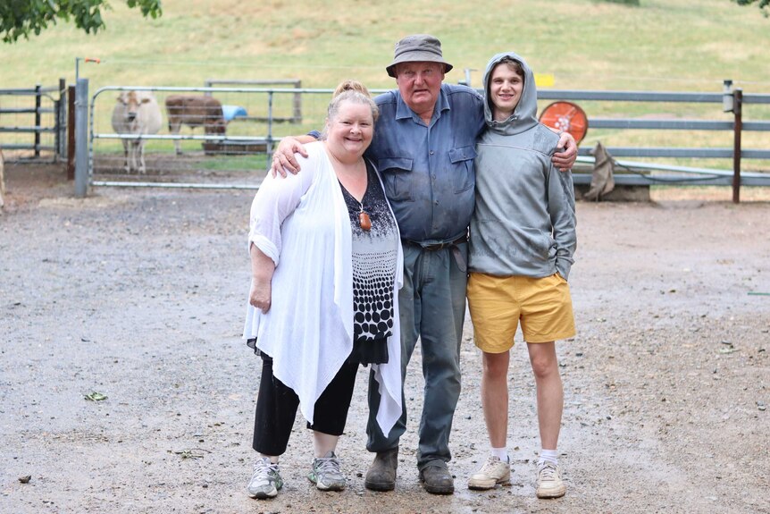An elderly farmer stands with comedian magda szubanski and a teenage boy wearing a hoodie, cattle in background