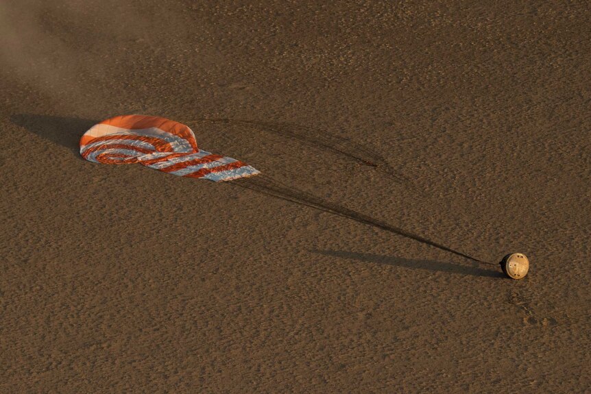 A spacecraft with its parachute deployed sits in the desert.