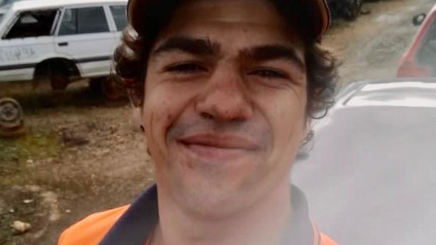 A smiling young man with dark, curly hair, wearing a cap and a high-vis vest. He is standing in a wrecking yard.