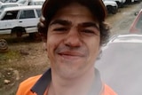 A smiling young man with dark, curly hair, wearing a cap and a high-vis vest. He is standing in a wrecking yard.