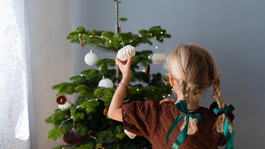 A behind view of a girl in blonde plaits decorating a minimalist Christmas tree