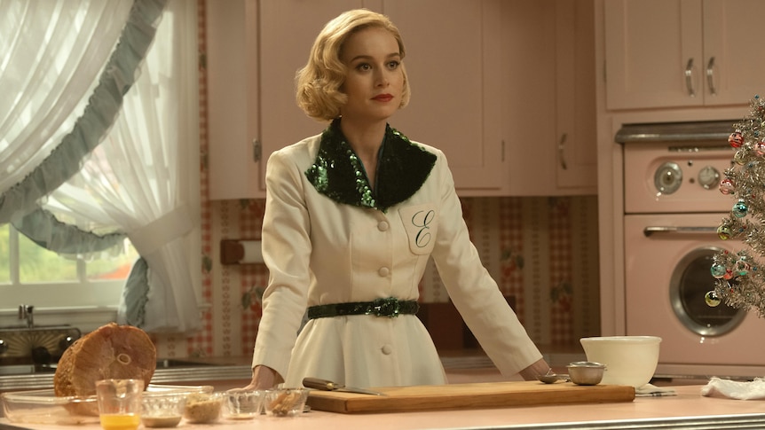 Actress Brie Larson with a 50s hairstyle and shiny green and white dress stands in a kitchen in the show Lessons in Chemistry.