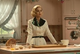 Actress Brie Larson with a 50s hairstyle and shiny green and white dress stands in a kitchen in the show Lessons in Chemistry.