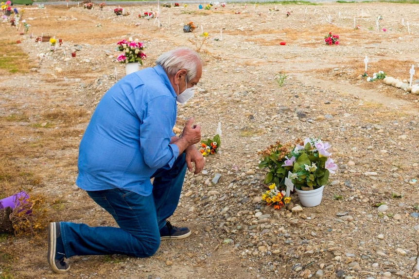 An older man in jeans and a shirt and face mask kneels before a small white cross in a field