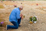 An older man in jeans and a shirt and face mask kneels before a small white cross in a field