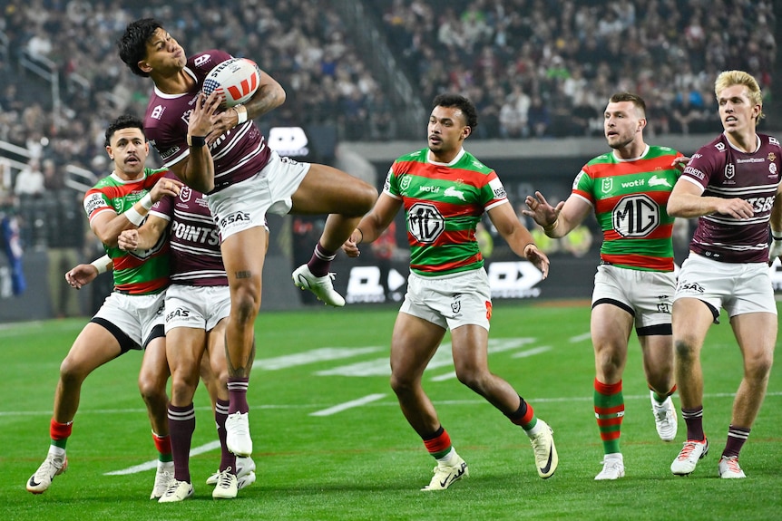 A Manly NRL player grasps the ball in both hands as he hangs in mid-air surrounded by players from both sides.