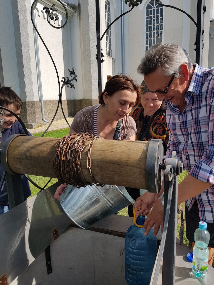 An older, grey-haired man smiles as a woman decants water from a rustic jug into a bottle.