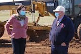 A woman in a pink blazer stands with hands on hips beside an older man wearing a hard hat in front of earthmoving equipment.
