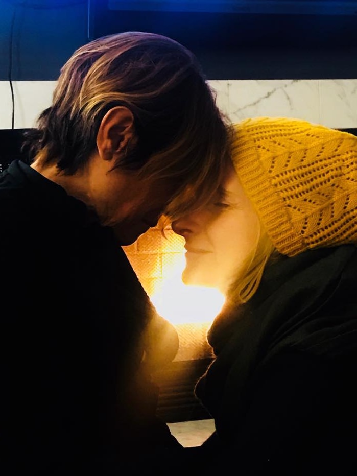 Two women nuzzle their heads close together in front of a fireplace. One is wearing a yellow beanie.