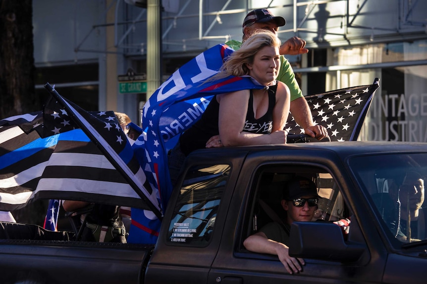 A woman wearing a Trump flag leans on the front of a ute as two men drive it.