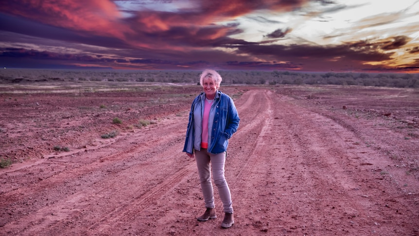 Woman stood smiling on a dirt road in the middle of a red dirt desert location. 