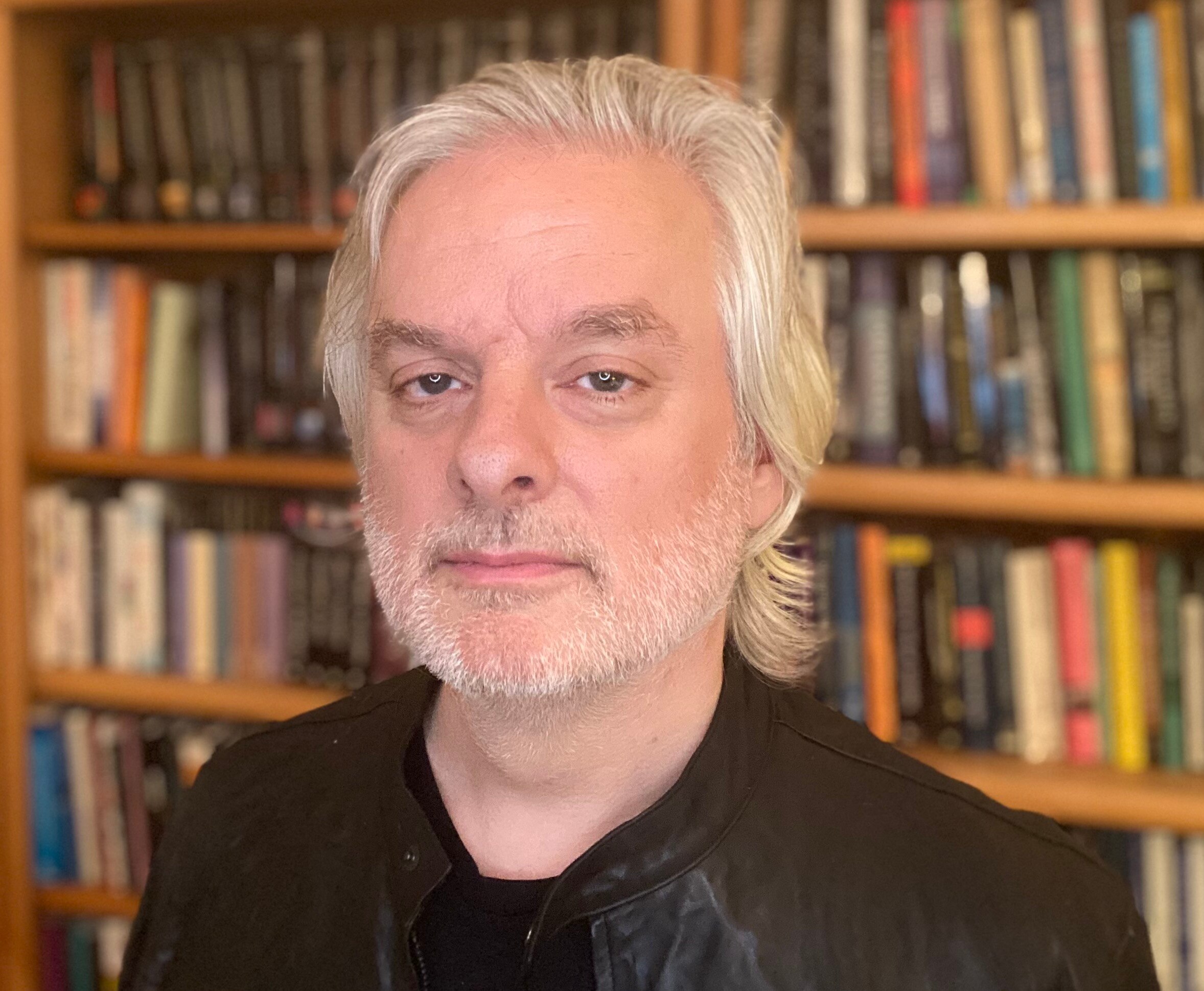 A man with straight, white hair and a neutral facial expression. A large bookcase is behind him.