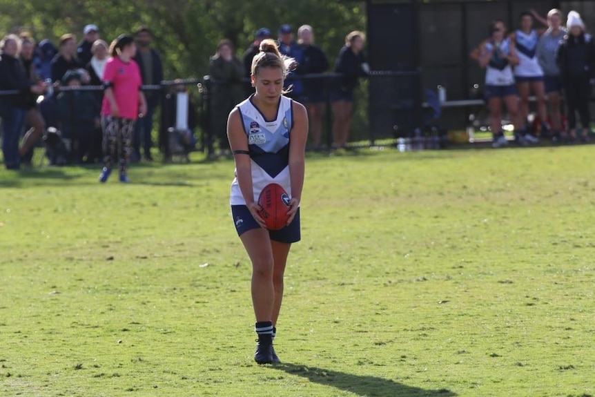 Ella Taverner stands on a sunny AFL field wearing a team singlet and shorts, and prepares to kick the AFL ball in her hands. 