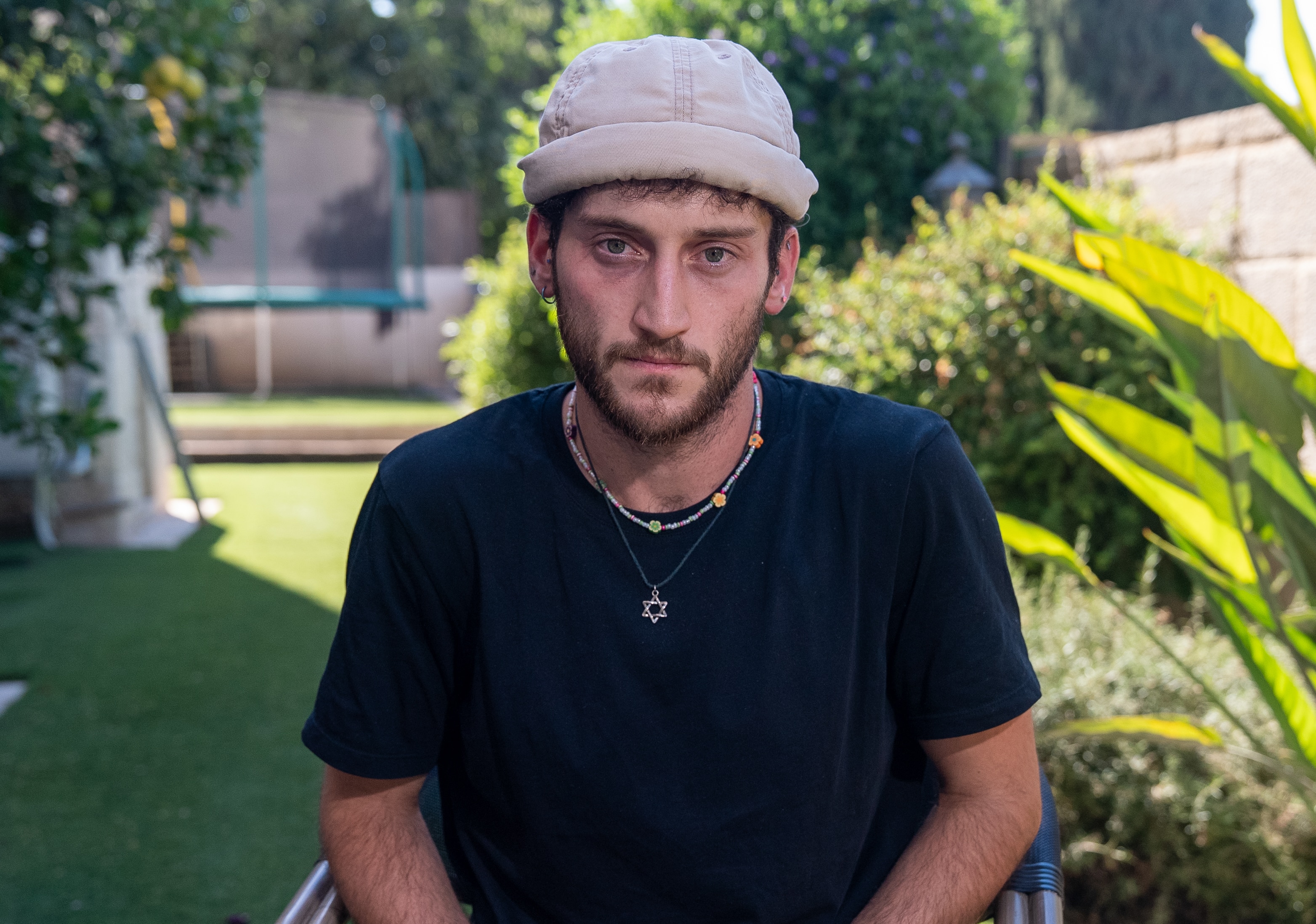 Yotam Vilk wears a hat, black t shirt and Star of David necklace as he sits in a garden and looks into the camera.