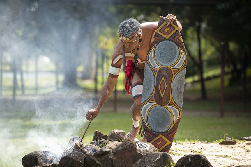 An Aboriginal main in tradition paint holds a decorated shield as he stokes a fire surrounded by rocks