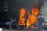 A man in a face mask running past a burning car. 