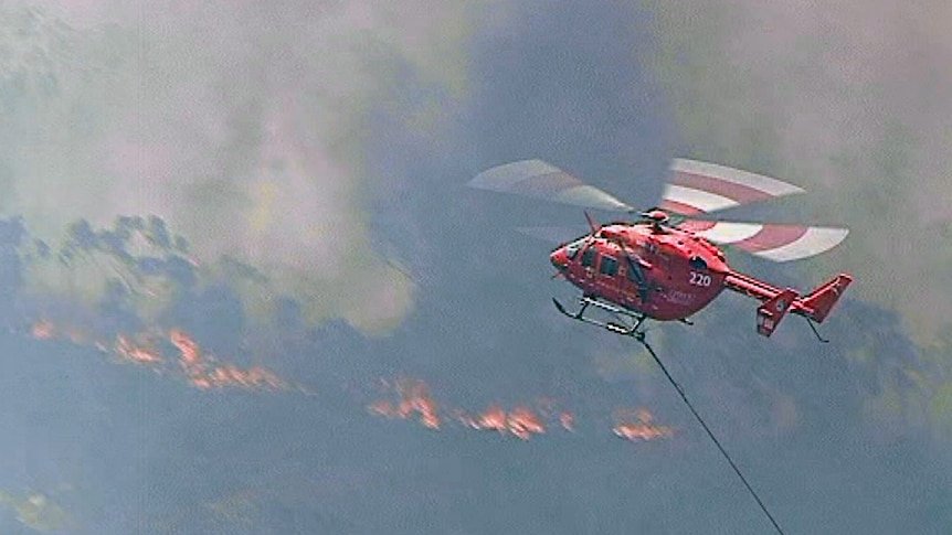 A red and white helicopter flies over burning bushland.