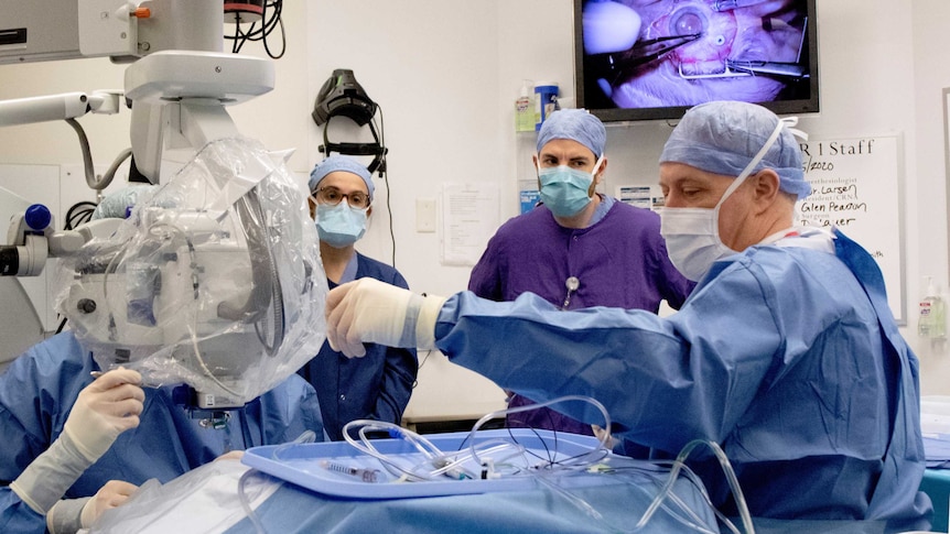 A doctor in blue hospital scrubs and face mask reaches for surgical equipment watched by colleagues.