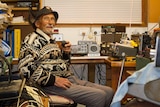 A old man sitting in front of a collection of radio equipment.