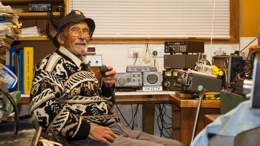 A old man sitting in front of a collection of radio equipment.