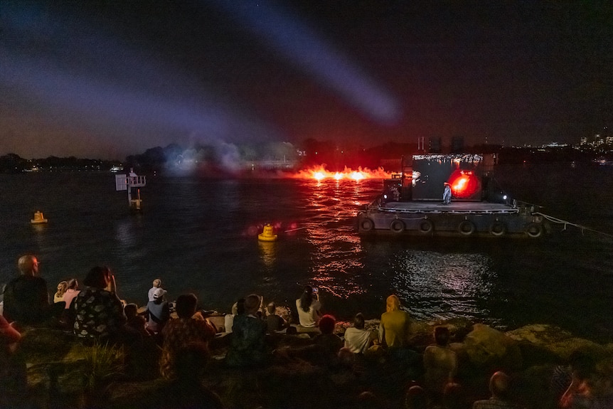 A First Nations woman stands on a barge, in front of a screen. Assembled on the rocks on shore is a group of people.