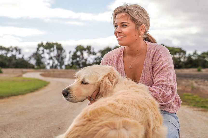 A girl kneels down by a golden retriever dog on a long country laneway.