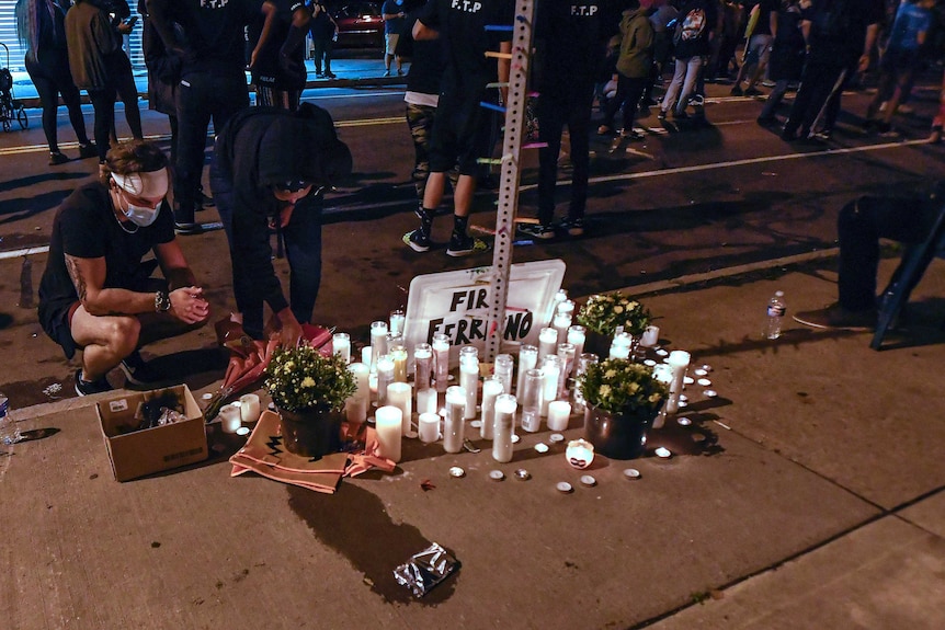 A group of people kneel and stand around a makeshift memorial with candles, flowers and signs.