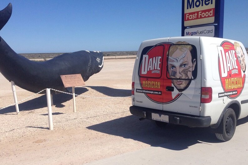 A statue of a whale next to a white Holden panel van covered in stickers with the face of magician Dane Certificate.