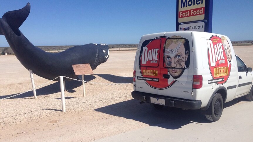A statue of a whale next to a white Holden panel van covered in stickers with the face of magician Dane Certificate.