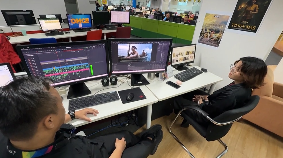 Two people sitting in front of the screens displaying a TV show scene.