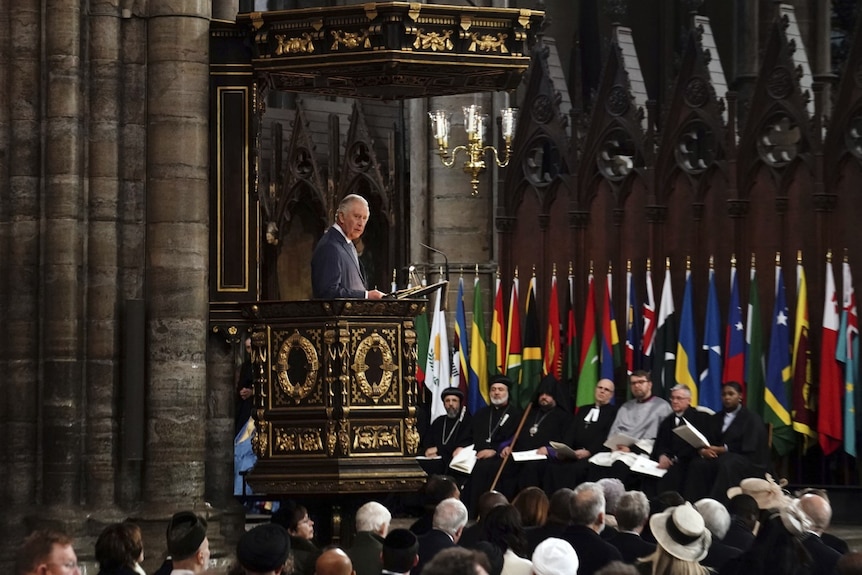 King Chalres stands on an ornate covered platform above a seated crowd inside the Abbey. On the back wall are an array of flags