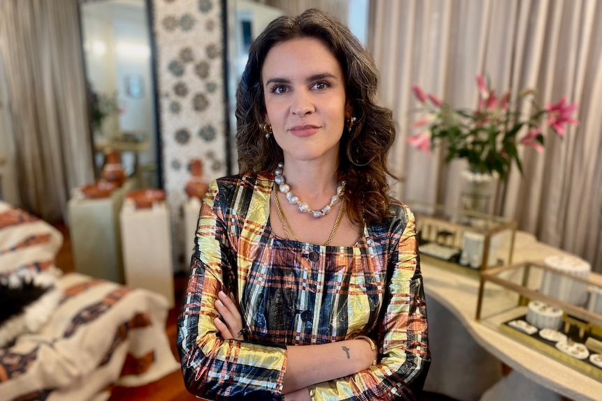 A female business owner in her thirties stands in her jewellery store and looks at the camera, arms folded 