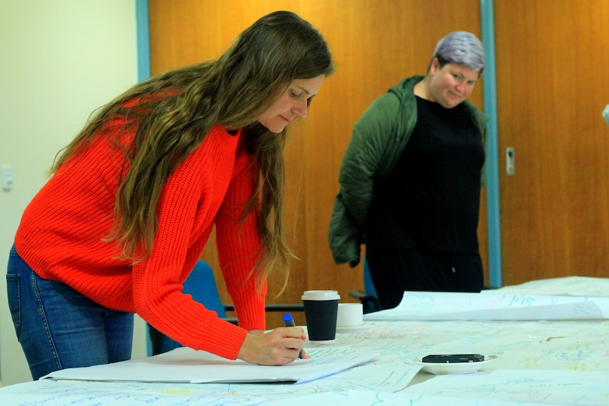 A woman in an orange jumper and long hair stands, leaning over a table to write