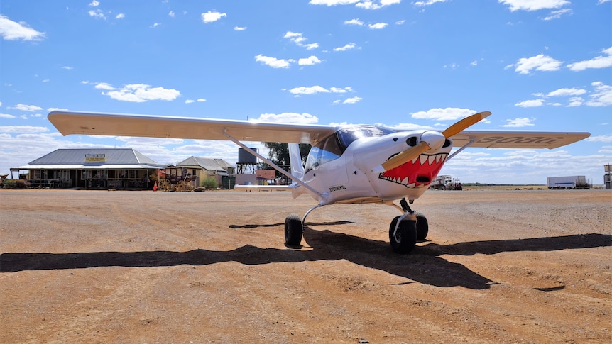 A small white plane with a jagged teeth painted on the front sits on a dirt road in front of an old outback pub.