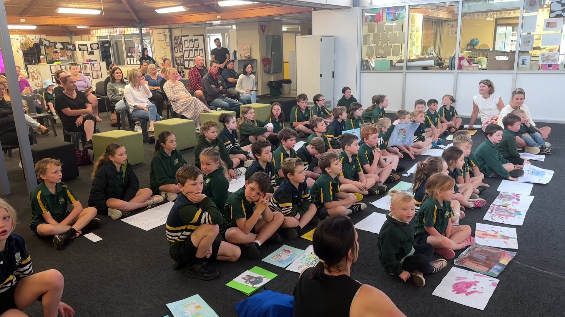 Group of children sitting on the floor with paper in front of them, adults sitting at back watch on.